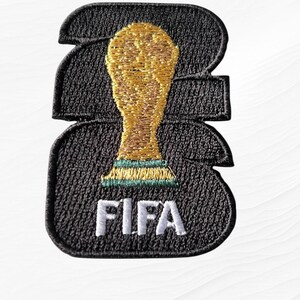 May include: A black embroidered patch featuring a gold-colored trophy icon centered on a stylized black number 22. The word FIFA is stitched in white thread at the bottom of the patch. The design has a thick black border and textured stitching.