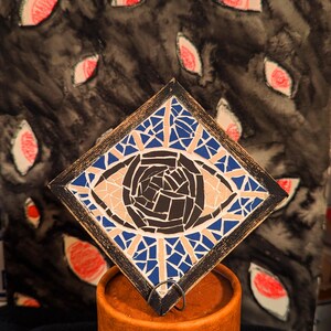 Peut inclure: Œuvre d'art représentant un œil en mosaïque sur un cadre carré, avec des carreaux bleus, beiges et noirs. L'œuvre est placée sur un fond sombre avec des éléments en forme d'œil rouges et blancs. Sur un récipient cylindrique orange.