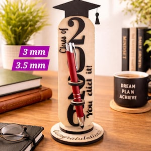 May include: A wooden graduation pen holder with a black mortarboard, the number 2026, and the text "Class of" and "You did it!". A red pen is inserted. The base reads "Congratulations!".