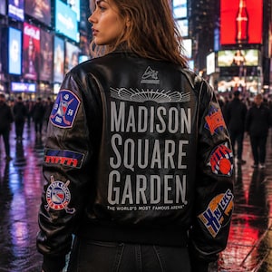 May include: A black leather varsity jacket featuring the text Madison Square Garden The World's Most Famous Arena on the back. The jacket includes various embroidered sports team patches on the sleeves and back, with a collar and ribbed cuffs.