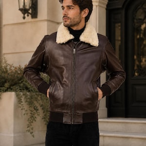 Puede incluir: Chaqueta bomber de cuero marrón con cuello de borrego color crema. La chaqueta tiene cremallera, puños de canalé y dobladillo de canalé. El modelo lleva pantalones negros.