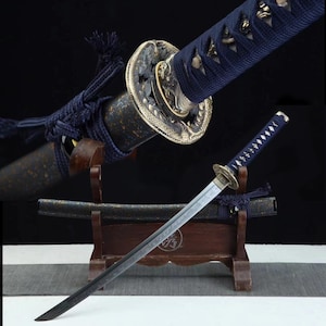 Puede incluir: Una katana japonesa con empuñadura y vaina azul oscuro. La hoja plateada se complementa con una guarda dorada con intrincados detalles. La espada descansa sobre un soporte de madera, mostrando su artesanía y diseño.