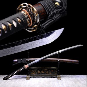 Può includere: Una spada katana giapponese con manico e lama neri. La lama ha una finitura argentata con un motivo unico. L'impugnatura è avvolta in un cordoncino nero e presenta dettagli dorati e argentati. La spada poggia su un supporto decorativo.