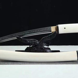 May include: A Japanese katana sword with a curved, black blade and a white handle. The sword rests on a black eagle-shaped stand, with a white scabbard below. The blade has a textured pattern.
