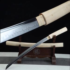 May include: A Japanese katana sword with a polished steel blade and a light-colored wooden handle and scabbard. The blade has a wavy pattern along the edge. The sword is displayed on a wooden stand.
