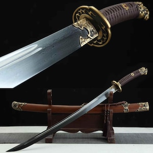 May include: A close-up of a sword featuring a brown handle and gold-toned decorative elements. The blade is silver with a dark pattern. The sword is presented in a wooden scabbard and stand, highlighting its intricate design.