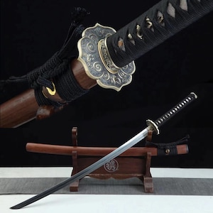 Puede incluir: Una espada katana japonesa con empuñadura y hoja negras. La espada presenta una guarda decorativa con un diseño ornamentado y una vaina de madera. El mango está envuelto en un cordón negro y la hoja es plateada.