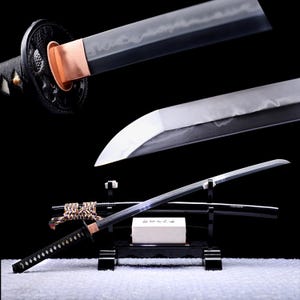 Puede incluir: Una espada katana japonesa con mango envuelto en negro, una abrazadera habaki de cobre y una hoja de acero con una línea hamon visible. La espada descansa sobre un soporte negro junto a su vaina negra, adornada con un cordón decorativo.