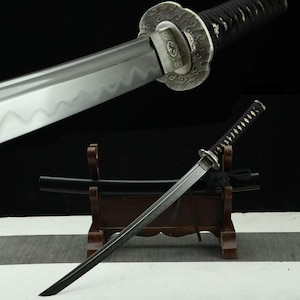 Op de afbeelding: Een katana zwaard met een zilverkleurig stalen lemmet met een golvende hamon-lijn, een zwart met koord omwikkeld handvat en een sierlijke, getextureerde zilverkleurige ronde stootplaat. Het zwaard wordt getoond met de bijpassende zwarte schede.