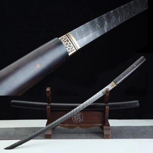 Op de afbeelding: Een Japanse katana-zwaard met een donkerhouten handvat en schede. Het lemmet heeft een patroon en een goudkleurige metalen bescherming. Het zwaard rust op een houten standaard.