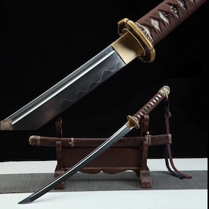 Pode incluir: Uma faca tanto japonesa com cabo marrom e guarda dourada. A lâmina é escura com um padrão ondulado. A faca é exibida em um suporte de madeira com uma bainha correspondente. Um pendão azul e vermelho pende da alça.
