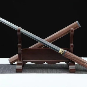 Könnte beinhalten: Ein japanisch anmutendes Schwert mit einer dunklen, strukturierten Klinge und einem Holzgriff. Das Schwert ruht auf einem Holzständer, mit einer passenden Scheide dahinter. Der Griff hat goldfarbene Akzente.