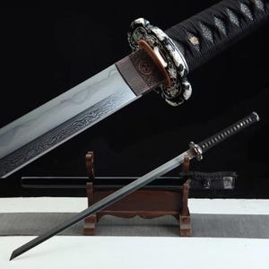 Puede incluir: Una espada katana japonesa con empuñadura negra y hoja plateada. La espada se exhibe sobre un soporte de madera, con una vaina negra visible. La hoja tiene un patrón único y el mango está envuelto en tela negra.