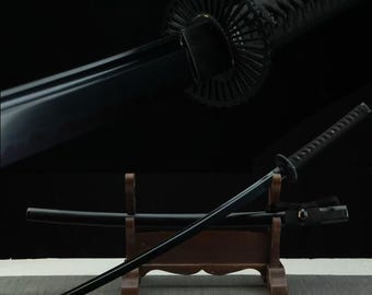 May include: A black katana sword with a matching black scabbard resting on a wooden stand. The sword features a dark blade, a circular tsuba guard with a radial pattern, and a handle wrapped in black cord. The design is entirely monochromatic.