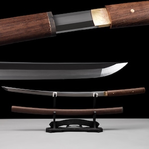 Może przedstawiać: Miecz katana z zakrzywionym stalowym ostrzem, ciemnobrązową drewnianą rękojeścią i pasującą drewnianą pochwą. Miecz spoczywa na czarnym stojaku, eksponując polerowaną metalową krawędź oraz teksturę drewna na rękojeści i pochwie.