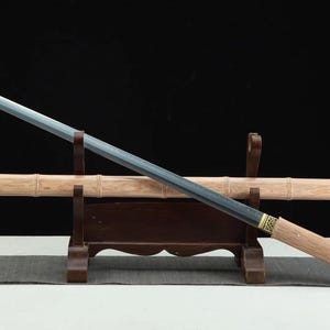 Puede incluir: Una espada de estilo japonés con una hoja recta gris y un mango de madera de color claro. La espada descansa sobre un soporte de madera marrón oscuro con una vaina de madera de color claro. El fondo es negro.