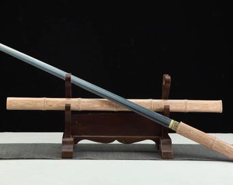 May include: A Japanese-style sword with a straight, gray blade and a light-colored wooden handle. The sword rests on a dark brown wooden stand with a light-colored wooden scabbard. The background is black.