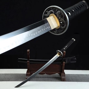 Pode incluir: Uma espada katana japonesa com cabo preto e lâmina prateada. A guarda apresenta um desenho de dragão. A espada é exibida em um suporte de madeira, com sua bainha visível.