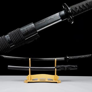 Può includere: Una spada katana nera con una lama curva, esposta su un supporto di legno. La spada presenta un'impugnatura strutturata e un fodero nero. La lama ha un motivo visibile.