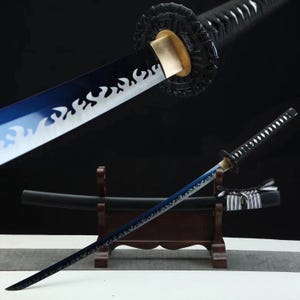 May include: A Japanese katana sword with a blue and white blade featuring a flame design. The sword has a black handle and is displayed on a wooden stand. The scabbard is black with a decorative ribbon.