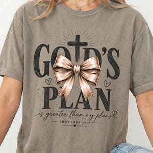 May include: A grey short-sleeved t-shirt featuring a graphic print with a large beige bow, a black cross, and the text "GOD'S PLAN is greater than my plans PROVERBS 16:9" in a stylised black font, accented with small hearts and starburst details.