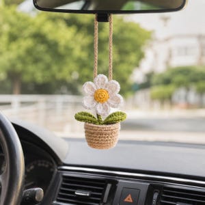 May include: A crocheted daisy car charm hanging from a rearview mirror. The white flower has a yellow center and green leaves, sitting in a tan crocheted pot. The charm is suspended by a tan cord, adding a touch of handmade charm to the car's interior.