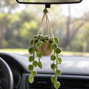 Puede incluir: Una planta colgante de ganchillo con una maceta marrón y hojas verdes, suspendida del espejo retrovisor de un coche. La planta está hecha de hilo y tiene un aspecto natural y hecho a mano. Las hojas caen hacia abajo, añadiendo un toque de naturaleza al interior del coche.