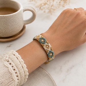 K&ouml;nnte beinhalten: Ein Armband mit Perlen in einem geometrischen Muster in Gold, T&uuml;rkis und Wei&szlig;. Das Armband wird am Handgelenk getragen und hat einen goldenen Kettenverschluss. Das Design umfasst kleine, perlenartige Perlen.