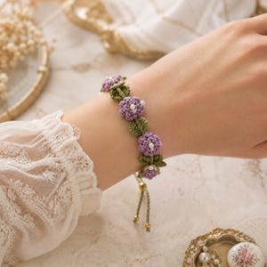 K&ouml;nnte beinhalten: Ein handgefertigtes Armband mit geh&auml;kelten lila Blumen mit wei&szlig;en Perlen in der Mitte und gr&uuml;nen Bl&auml;ttern. Das Armband hat eine hellgr&uuml;ne Kordel mit goldfarbenen Akzenten. Das Armband wird am Handgelenk getragen.