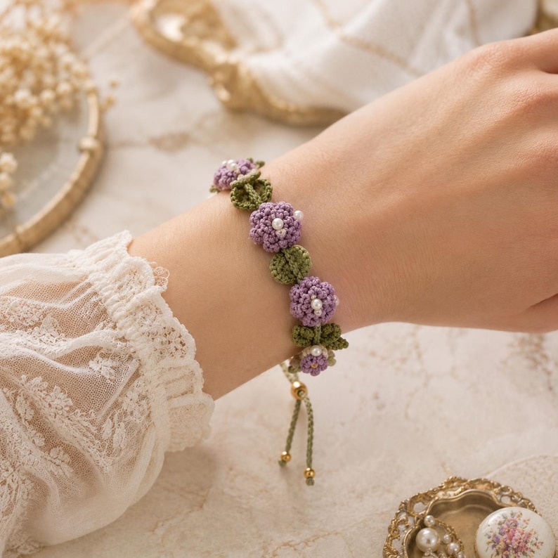 K&ouml;nnte beinhalten: Ein handgefertigtes Armband mit geh&auml;kelten lila Blumen mit wei&szlig;en Perlen in der Mitte und gr&uuml;nen Bl&auml;ttern. Das Armband hat eine hellgr&uuml;ne Kordel mit goldfarbenen Akzenten. Das Armband wird am Handgelenk getragen.