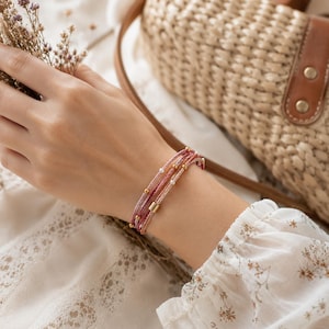 K&ouml;nnte beinhalten: Ein mehrstr&auml;ngiges Armband in Rosa- und Cremet&ouml;nen, verziert mit kleinen goldenen Perlen und rechteckigen Akzenten. Das Armband wird am Handgelenk getragen, mit einer Strohtasche und floralen Details im Hintergrund.