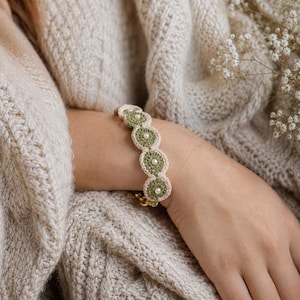 K&ouml;nnte beinhalten: Ein handgefertigtes Armband mit einer Reihe von gr&uuml;nen und cremefarbenen geh&auml;kelten Kreisen, die jeweils mit einer kleinen Perle verziert sind. Das Armband wird mit einem goldfarbenen Verschluss befestigt. Das Armband wird am Arm einer Person gezeigt.