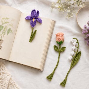 以下が含まれることがあります： かぎ針編みの花のブックマーク。紫のアイリス、桃色のバラ、白いスズランの形です。各ブックマークには緑色の茎とタッセルが付いています。ブックマークは開いた本の上に置かれています。