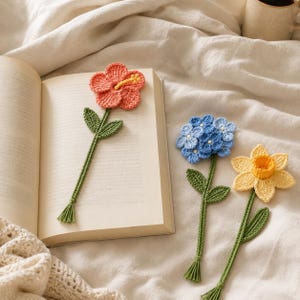 May include: Crocheted flower bookmarks in coral, blue, and yellow, with green stems and leaves. The bookmarks are placed in an open book on a cream-colored surface. The coral flower has a yellow center.