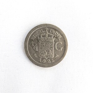 Puede incluir: Una moneda de plata de 1921 con la inscripción "NEDERL. INDIE" y "1/4 G". La moneda presenta una corona sobre un escudo de armas. La moneda circular tiene un aspecto ligeramente desgastado.