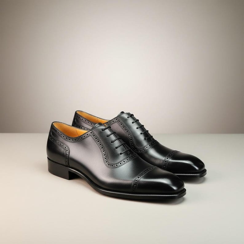 May include: A pair of black leather dress shoes with a cap toe design. The shoes have a classic Oxford style with intricate stitching and a lace-up closure. The shoes are displayed on a neutral background.