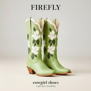 Puede incluir: Botas de vaquera verde claro con diseños florales blancos y hojas verdes oscuras. Las botas tienen punta, tacón de madera y borde superior festoneado con ribete rosa y blanco. La palabra "FIREFLY" está en la parte superior y "cowgirl shoes crafted for durability" en la parte inferior.