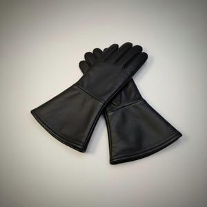 Puede incluir: Un par de guantes de cuero negro con puños acampanados. Los guantes están hechos de cuero liso y tienen un diseño clásico. Los guantes son probablemente para disfraces o recreaciones históricas.