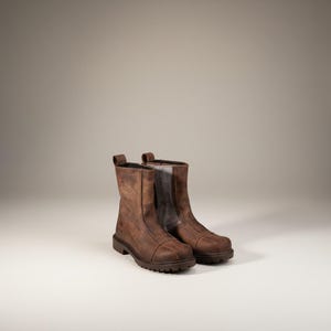Könnte beinhalten: Ein Paar braune Lederstiefel mit einem robusten, abgenutzten Aussehen. Die Stiefel haben eine mittlere Wadenhöhe, eine runde Spitze und eine robuste Sohle. Die Stiefel sind etwa 20 cm hoch.