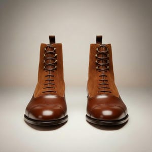 May include: A pair of brown leather and suede boots with a cap toe design. The boots have a lace-up closure with brown laces and a pull tab at the top. The boots are displayed on a white background.