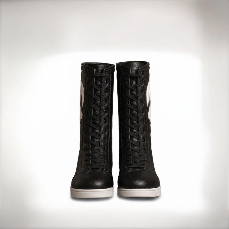 K&ouml;nnte beinhalten: Ein Paar schwarze wadenhohe Stiefel mit durchgehender Schn&uuml;rung vorne, schwarzen Schn&uuml;rsenkeln, einer strukturierten lederartigen Oberfl&auml;che und einer massiven wei&szlig;en Gummisohle. Ein gro&szlig;es wei&szlig;es kreisf&ouml;rmiges Logo ist an der Au&szlig;enseite.