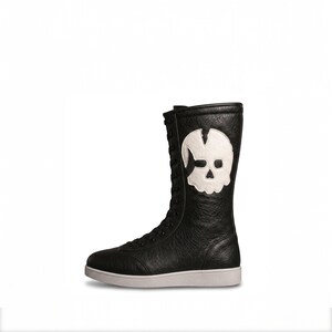 K&ouml;nnte beinhalten: Ein schwarzer, wadenhoher Schn&uuml;rstiefel mit strukturierter Oberfl&auml;che. An der Seite des Stiefels befindet sich eine gro&szlig;e, wei&szlig;e, aufgen&auml;hte Totenkopf-Applikation. Der Stiefel hat eine flache, durchgehende wei&szlig;e Gummisohle und schwarze Schn&uuml;rsenkel.