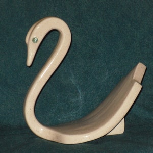 May include: A ceramic swan figurine with a light pink glaze and green glass eyes. The swan is in a graceful pose with its neck curved and its wings spread out.