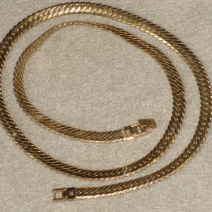 May include: A gold-colored necklace with a herringbone chain design. The necklace is laid out in a circular pattern, with a clasp visible at the bottom. The chain has a textured surface and a shiny finish.