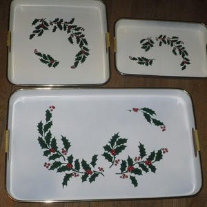 May include: A set of three white serving trays with gold-toned rims and yellow-textured side handles. Each tray features a central graphic of green holly leaves and red berries arranged in a curved pattern across the smooth white surface.