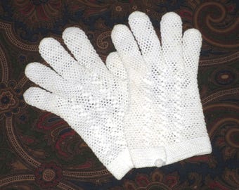 May include: A pair of white, open-weave crochet gloves. The gloves are made of a delicate, lacy material, with a button closure at the wrist. The gloves are displayed on a patterned background.
