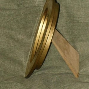 Puede incluir: Un marco de fotos redondo de color dorado con un soporte de madera. El marco tiene un borde elevado y un acabado liso.