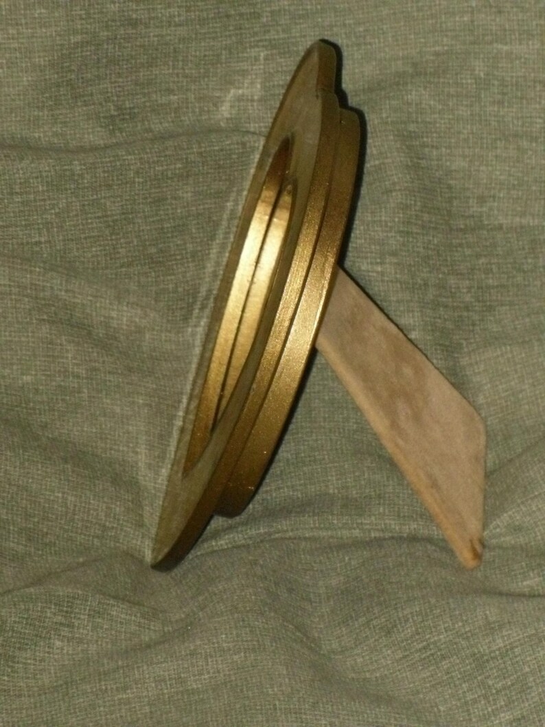 Puede incluir: Un marco de fotos redondo de color dorado con un soporte de madera. El marco tiene un borde elevado y un acabado liso.