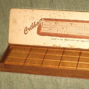 May include: An open vintage cribbage board game box. The box is brown with a cream-colored lid featuring the word "Cribbage" and a diagram of the board. The box contains gold-colored pegs and the text "THERE IS NO SUBSTITUTE FOR QUALITY".