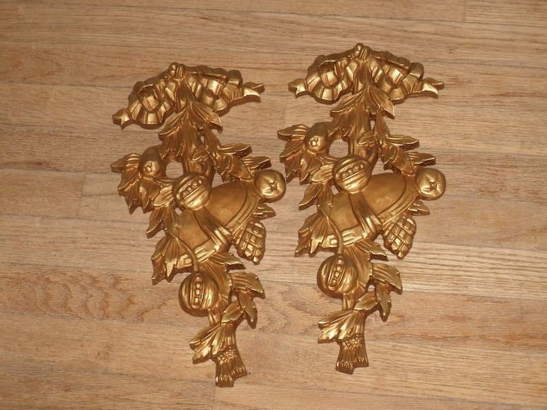 May include: A pair of gold-colored metal wall decor pieces featuring a stylized design of bells, leaves, and berries.